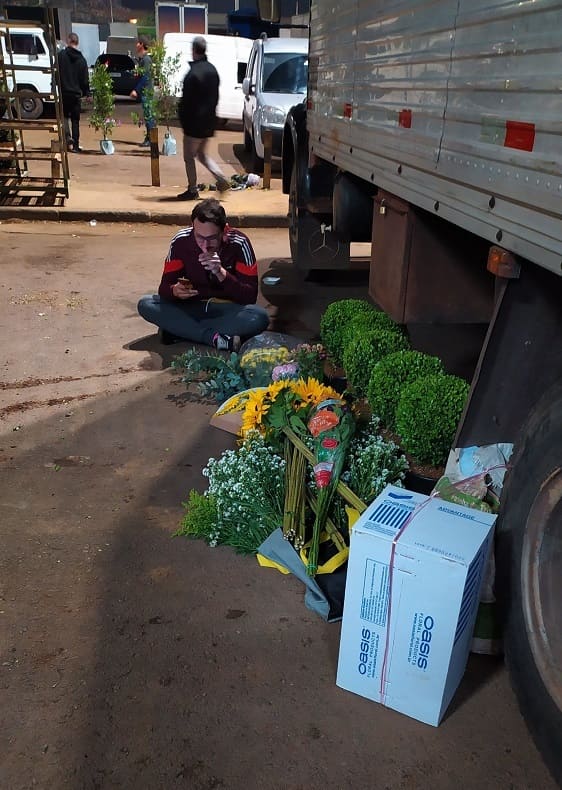 Homem sentado ao fundo, ao lado de um caminhão na Feira de Flores do CEAGESP, com flores e plantas no chão à sua frente