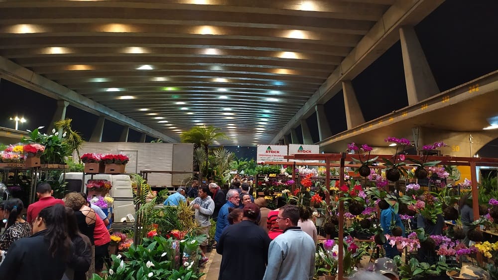 Foto da Feira de Flores do CEAGESP: um galpão comprido, com flores aos lados e muitas pessoas passando no centro