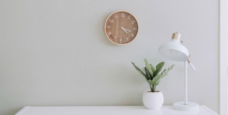 Parede branca com um relógio, um vaso de plantas e uma luminária sobre uma mesa, capa do texto sobre o que fazer na quarentena