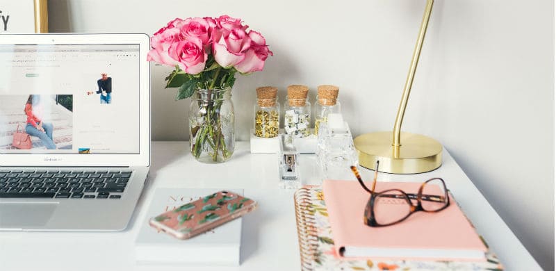 Imagem de um home office, com um computador, um pote com flores, cadernos e um óculos. Estudar na quarentena