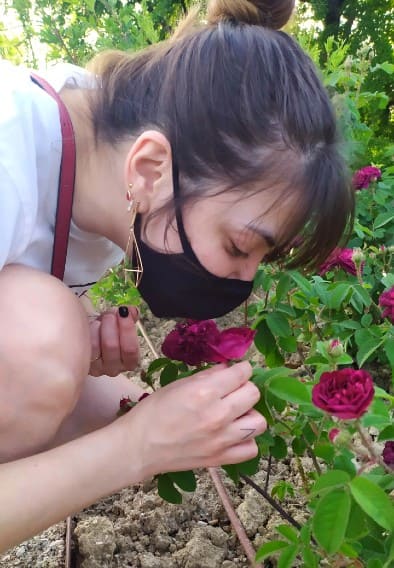 Moça de máscara preta cheirando uma flor roxa no Roseto di Santa Giustina, em Pádua