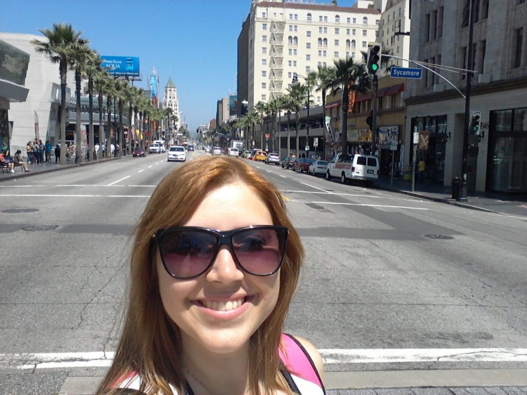 Selfie feita no meio da rua em Hollywood Boulevard, em Los Angeles. A rua está vazia, com alguns carros estacionados e algumas pessoas nas calçadas.