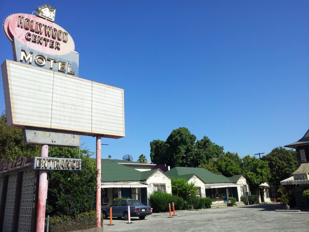 Fachada do motel Hollywood Center, com um letreiro descascado e uma casa ao fundo, parecendo abandonado